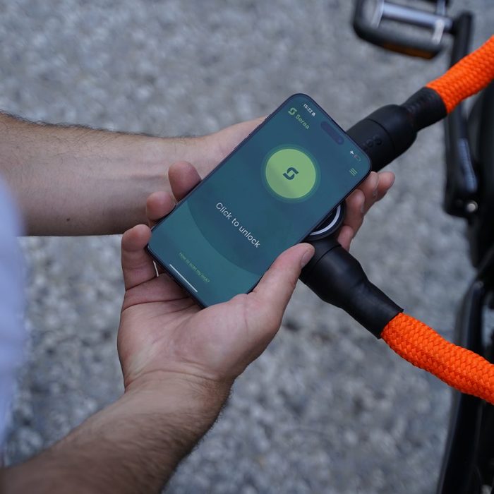 Het scherm van de Serea-app vraagt de gebruiker om met een klik het slot te ontgrendelen. Daarachter bevindt zich het Serea-slot dat tegen de mobiele telefoon wordt gehouden.